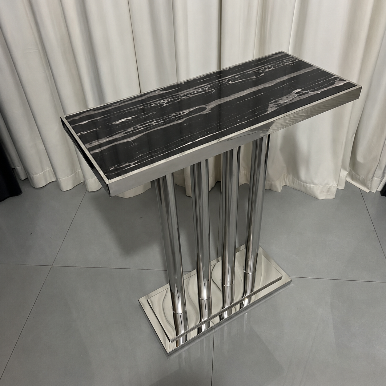 A modern rectangular console table with a glossy black and white marble-patterned top and a polished silver metal pedestal base, set against a background of white curtains and gray tiled flooring.