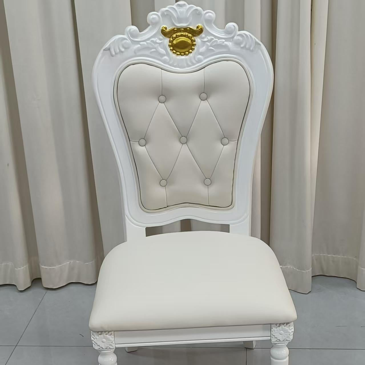 An ornate white dining chair with a tufted cream upholstered backrest and seat, featuring a decorative gold crest on the top of the backrest, set against a background of beige curtains and a gray tiled floor.