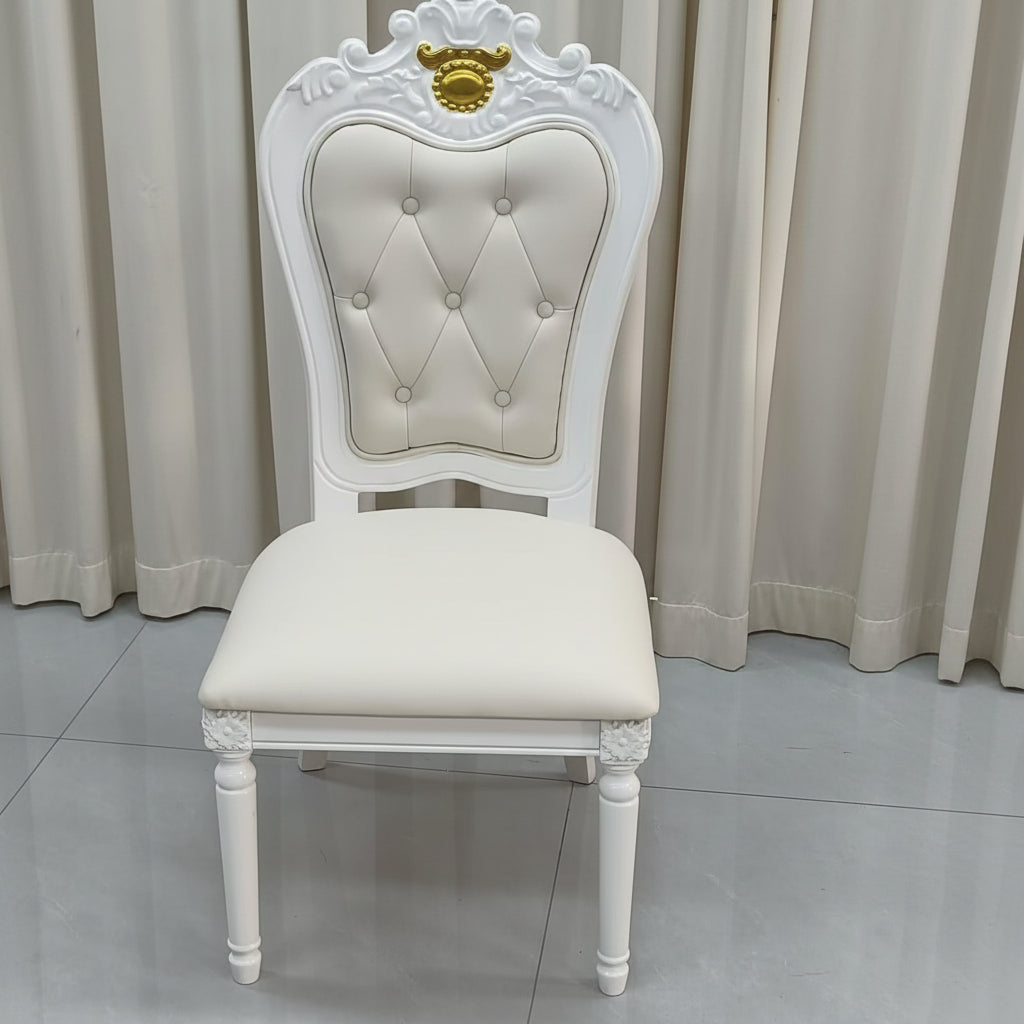 A white ornate dining chair with a tufted cream upholstered backrest and seat, featuring a gold crest detail on the top of the backrest, set against a neutral curtain and tiled floor background.