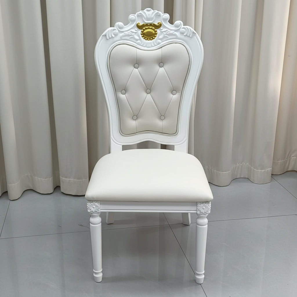 An ornate white baroque-style dining chair with a tufted cream upholstered backrest and seat cushion, featuring a gold crest detail on the top of the backrest, set against a background of beige curtains and a gray tiled floor.