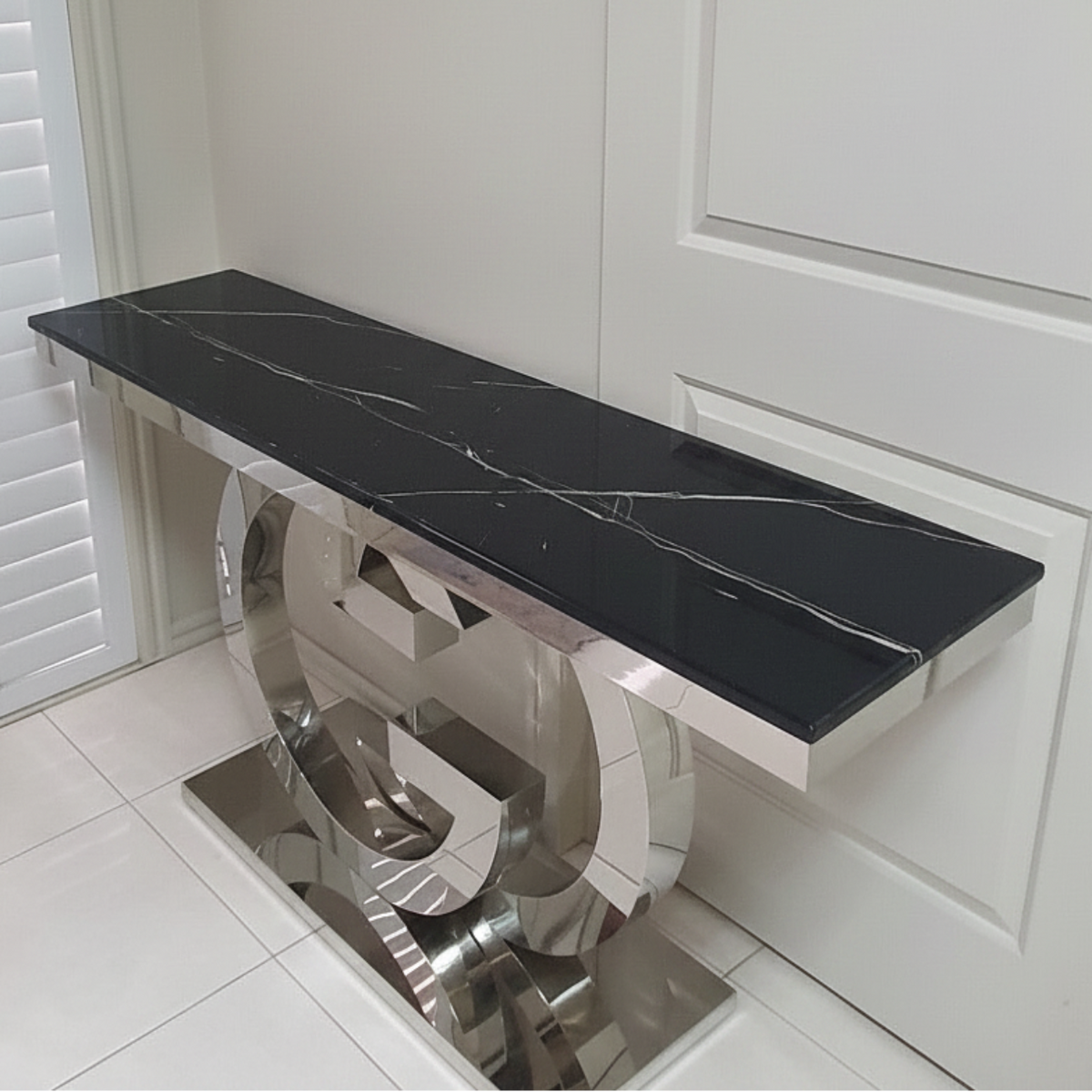 A modern console table with a glossy black marble top featuring white veining and a polished chrome base, positioned against a white paneled wall.