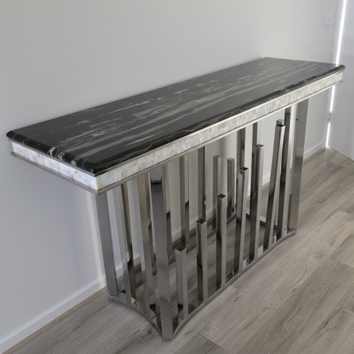 A modern console table with a dark gray wood top and a polished silver metal base featuring vertical slats and angled legs, shown in a room with light gray walls and wood-look flooring.