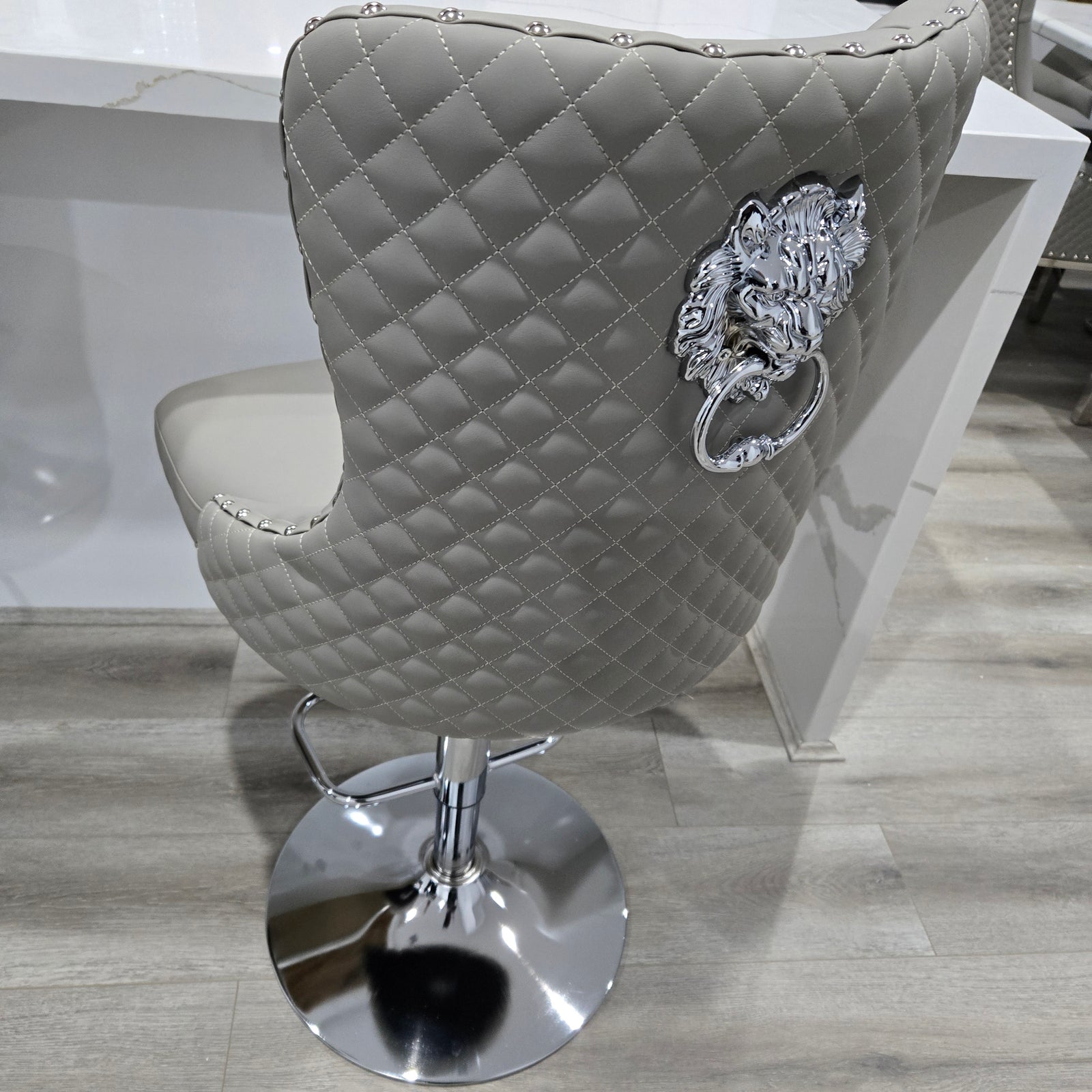 Grey Leather quilted bar stool with gold base and lion emblem against a white curtain background