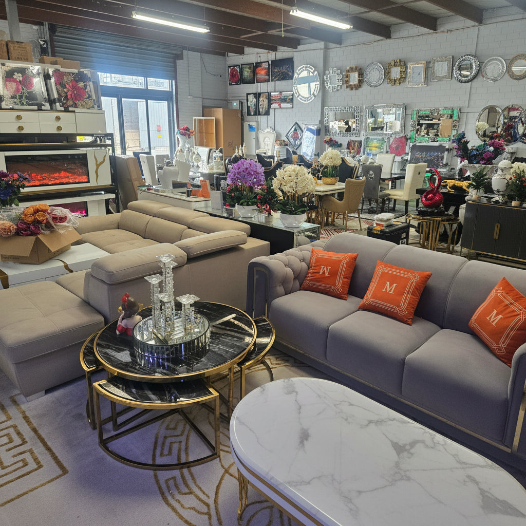 A modern living room furniture showroom featuring a gray tufted sofa with orange accent pillows, a beige sectional sofa, and a round black and gold coffee table in a busy retail environment.