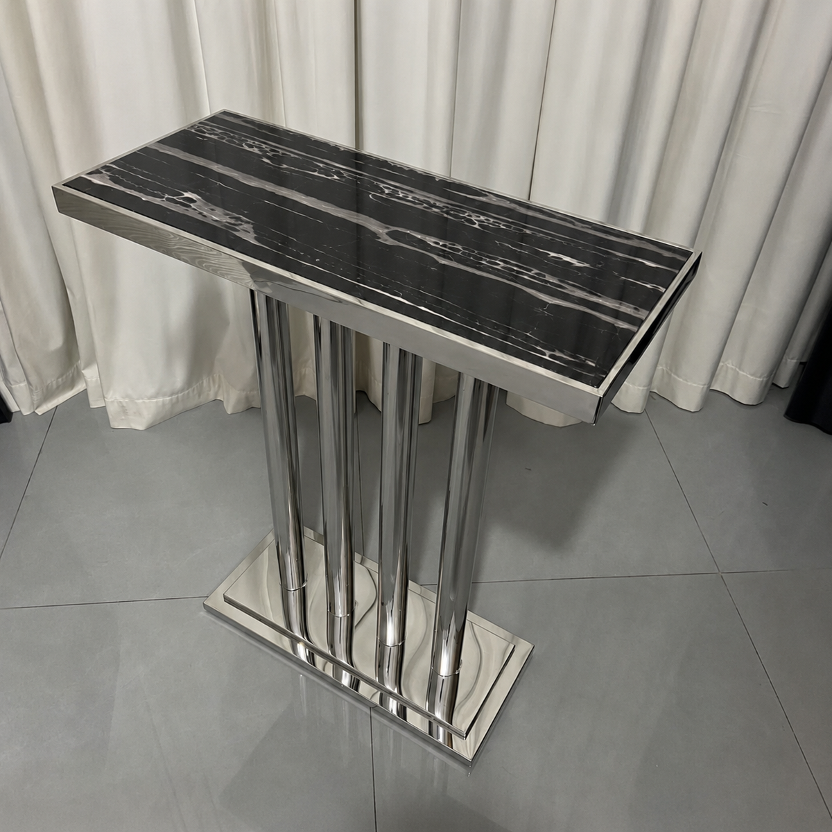 A modern rectangular console table with a glossy black and white marble-patterned top and a polished silver metal pedestal base, set against a background of white curtains and gray tiled flooring.
