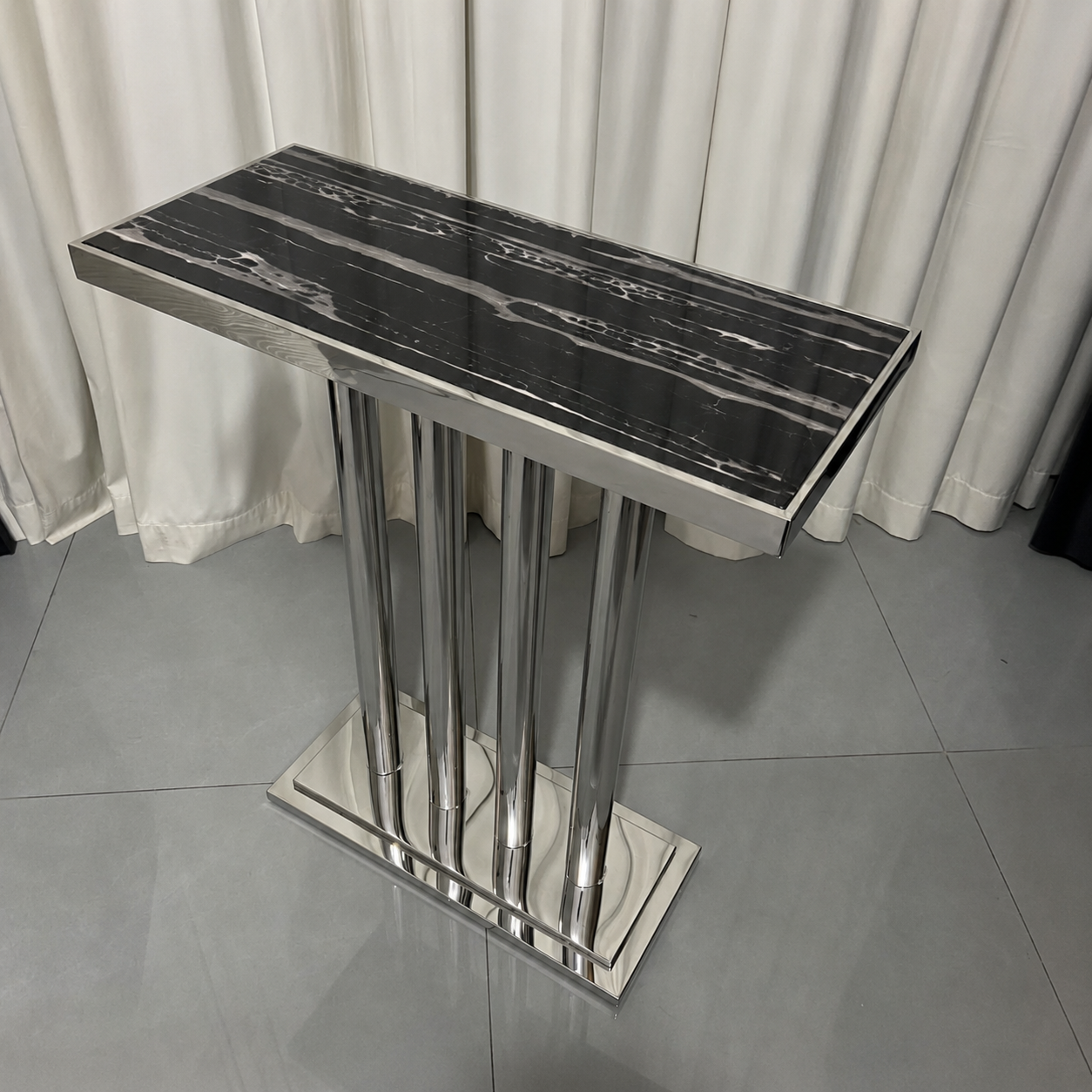 A modern rectangular console table with a glossy black and white marble-patterned top and a polished silver metal pedestal base, set against a background of white curtains and gray tiled flooring.