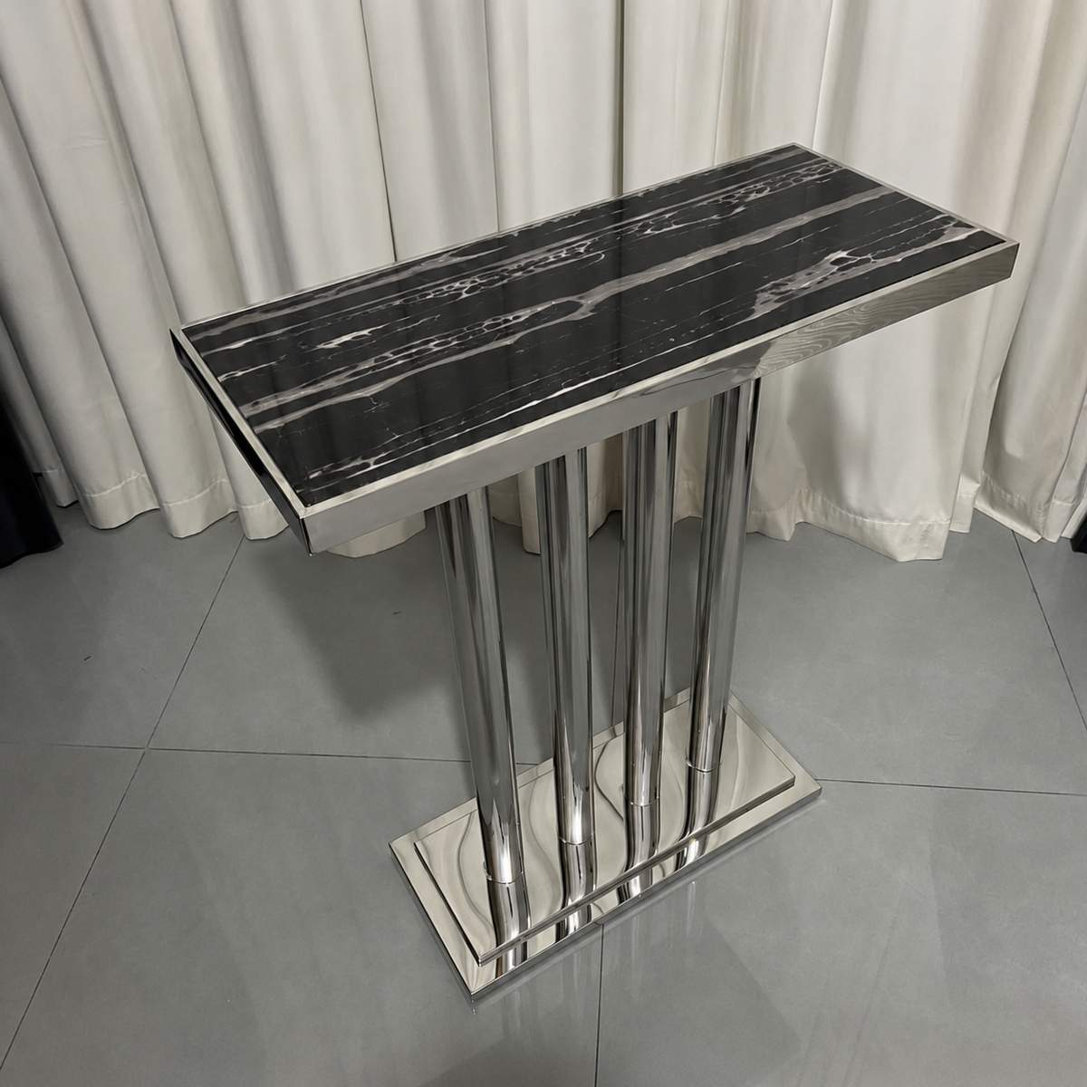 A modern rectangular console table with a glossy black and white marble-patterned top and a polished silver metal pedestal base, set against a background of white curtains and gray tiled flooring.