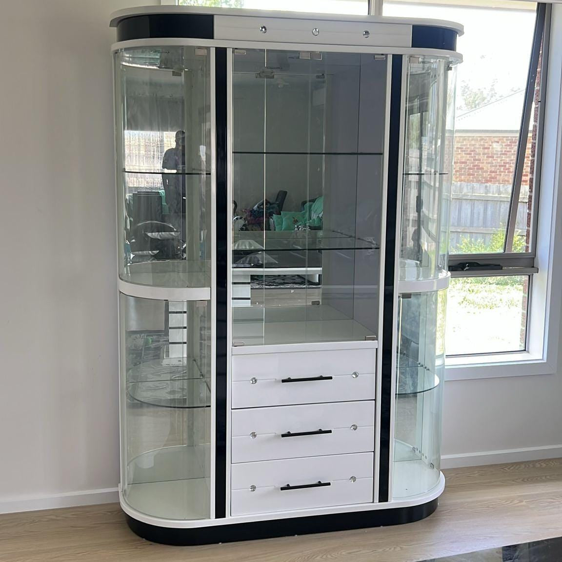 Curved glass display cabinet with white base and black accents in a room with a window.