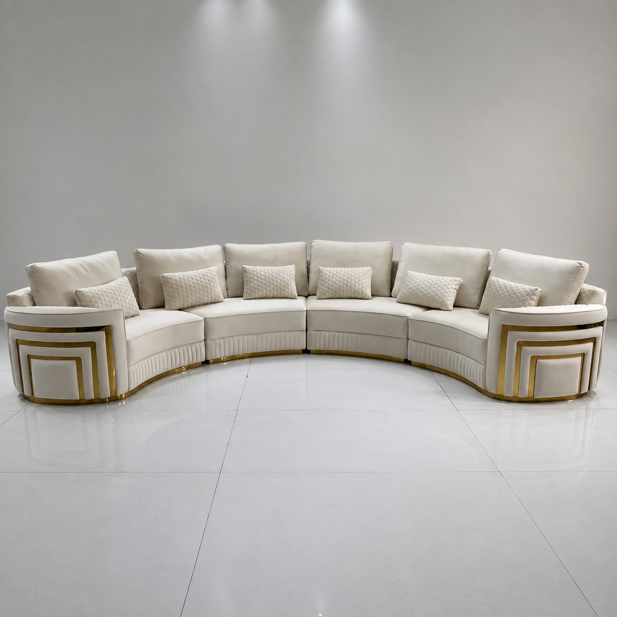 A curved modular sectional sofa upholstered in warm off-white fabric with decorative gold geometric trim on the outer arms and base, shown in a minimalist studio setting with a light gray wall and white floor.