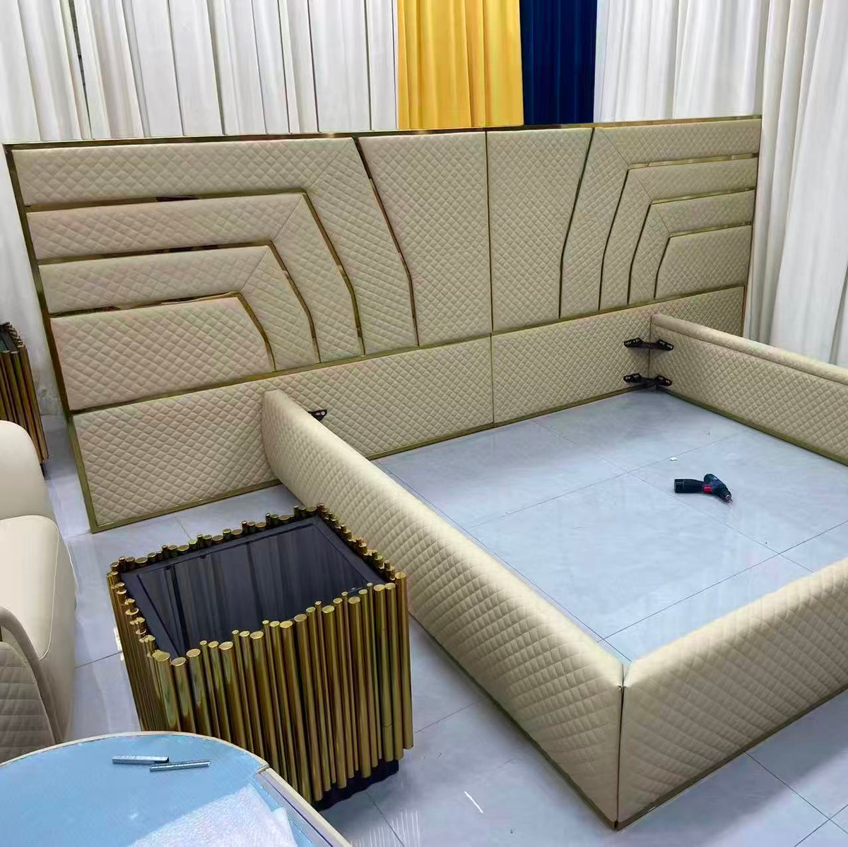 Beige upholstered bed frame in a room with curtains and a side table.