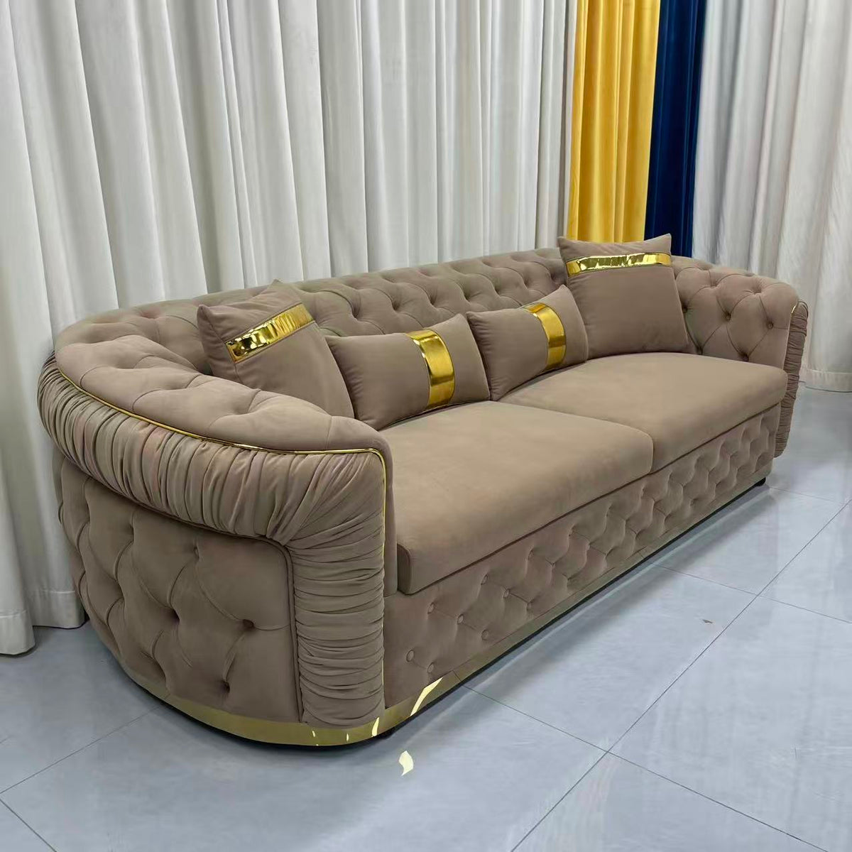 Beige sofa with gold accents in a room with white curtains and a tiled floor.