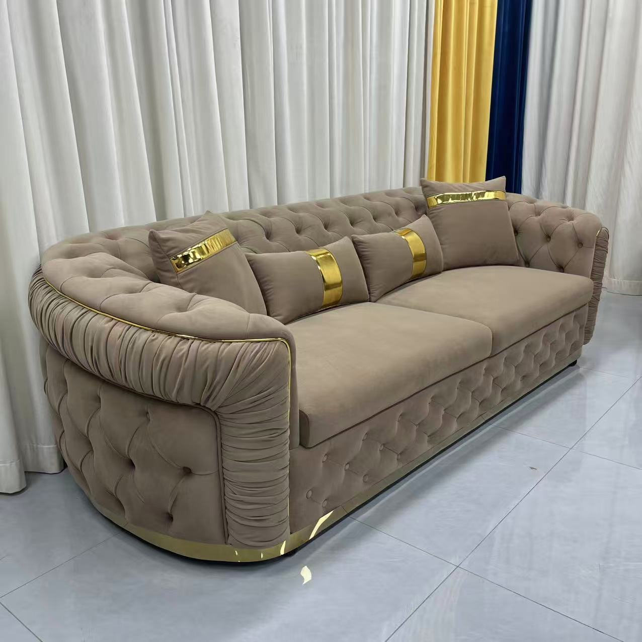 Beige sofa with gold accents in a room with white curtains and a tiled floor.