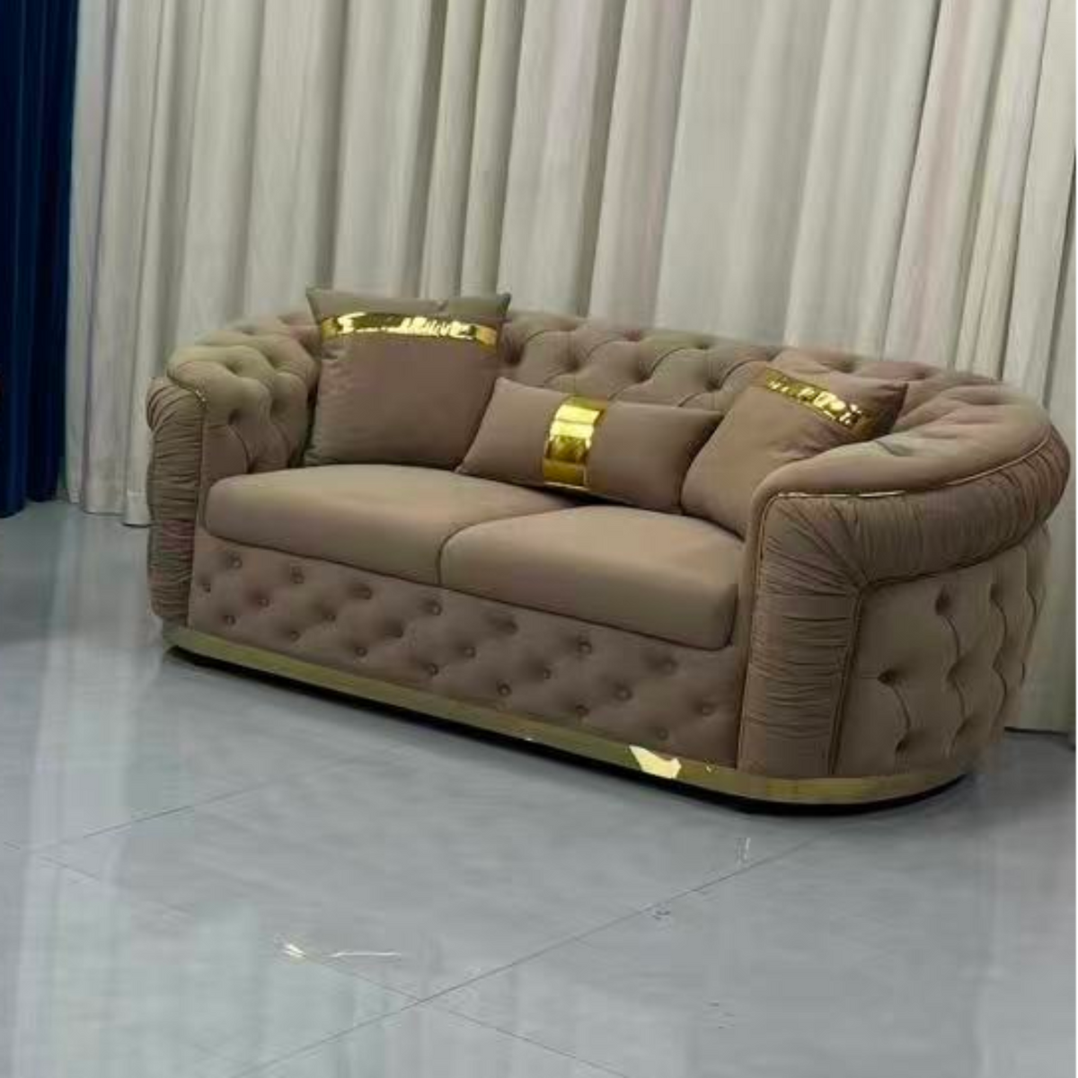 Beige tufted sofa with gold accents in a room with white curtains.