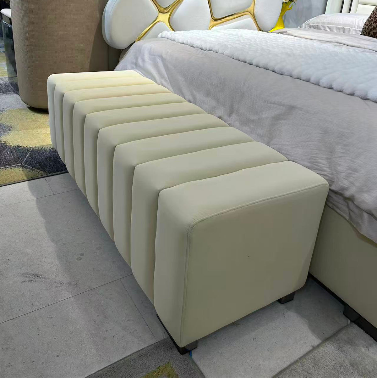 Beige ottoman in front of a bed with a modern headboard in a bedroom setting.