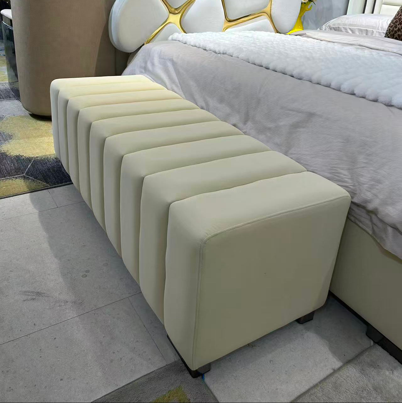 Beige ottoman in front of a bed with a modern headboard in a bedroom setting.