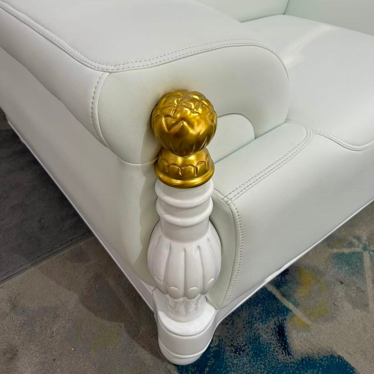 Close-up of a white leather armchair with a tufted backrest and ornate gold-and-white turned wooden armrests, shown in an indoor setting on a patterned rug.